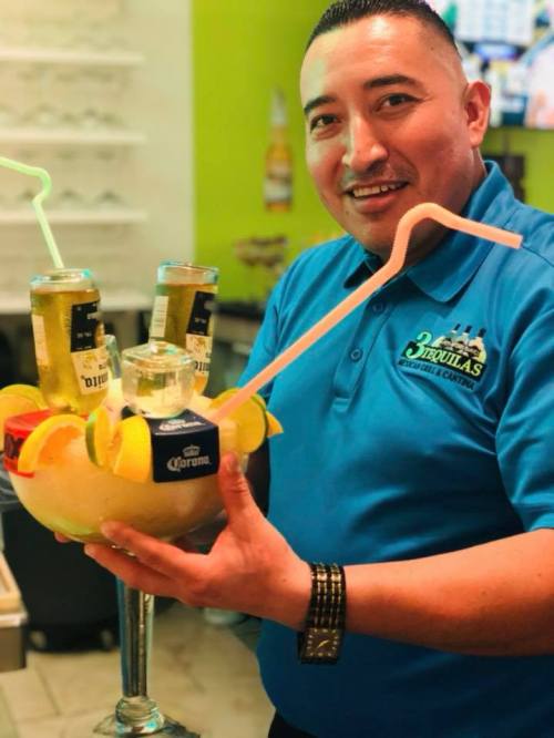 A smiling man in a blue shirt holds a large drink with beer bottles and fruit garnishes, set against a vibrant background.