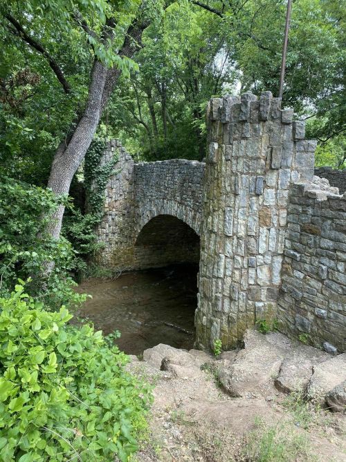 A stone bridge with an arch over a small stream, surrounded by lush greenery and trees.