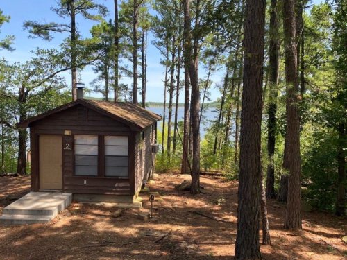 A small brown cabin surrounded by trees, overlooking a serene lake under a clear blue sky.