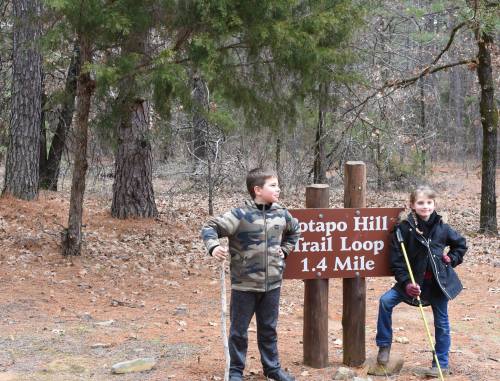 Two children pose playfully beside a sign for the Potapo Hill Trail Loop, surrounded by trees and fallen leaves.