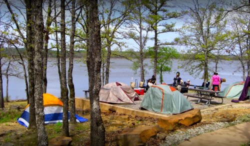 A lakeside campsite with tents, picnic tables, and people enjoying the outdoors among trees and water.