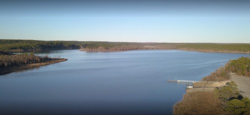 A serene lake surrounded by trees, with a small dock and parking area visible along the shore. Clear blue sky above.