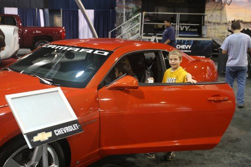 A young boy stands by the open door of a red Chevrolet Camaro at an auto show, smiling at the camera.