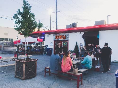 A lively outdoor scene with people socializing at tables near a food establishment, with an "OPEN" sign visible.