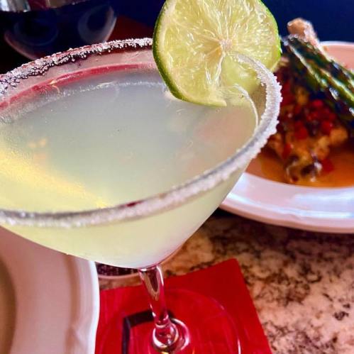 A close-up of a margarita glass with a lime wedge and salt rim, next to a plate of food.
