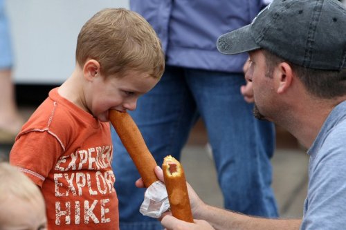 A child and an adult share a fun moment, both holding corn dogs, with the child playfully biting one.