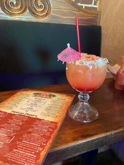 A colorful cocktail with a small umbrella and coconut flakes, next to a menu on a wooden table.