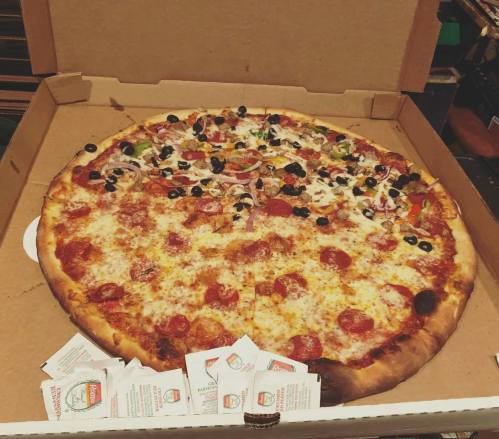 A large pizza with pepperoni, olives, and vegetables in a pizza box, surrounded by dipping sauce packets.