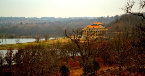 A scenic view of a large house with an orange roof, surrounded by trees and a calm lake in a rural landscape.