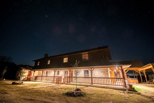 A well-lit house at night, surrounded by a starry sky and a grassy yard.
