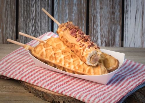 A white tray holds three waffle corndogs topped with bacon bits, resting on a red-striped cloth.