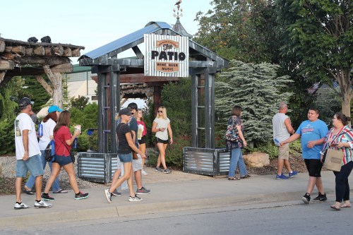 A lively outdoor patio entrance with people walking by, surrounded by greenery and rustic decor.
