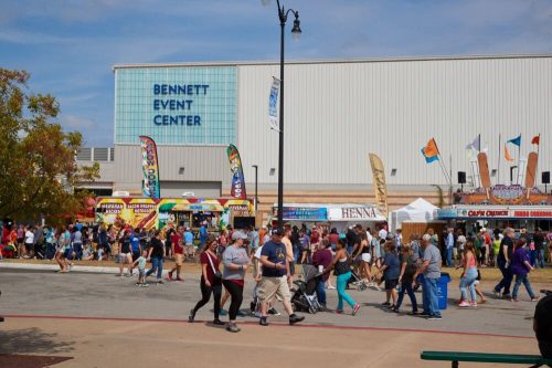 Crowd of people walking outside the Bennett Event Center, with food stalls and colorful banners in the background.