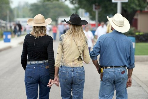 Three people in cowboy hats and denim walking together at an outdoor event, viewed from behind.