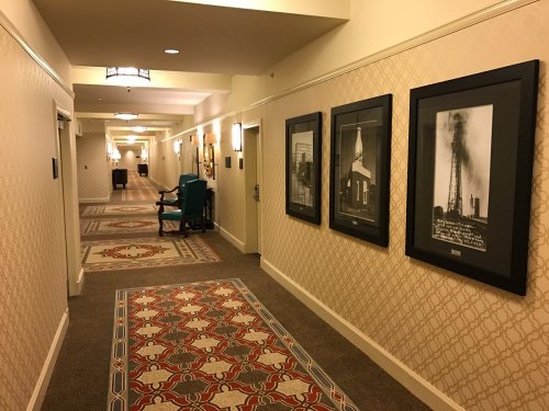 A hotel hallway with patterned carpet, framed black-and-white photos on the walls, and a green chair in the corner.