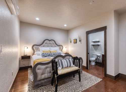Cozy bedroom with a plush bed, decorative bench, and an adjoining bathroom. Warm lighting and neutral tones create a welcoming atmosphere.
