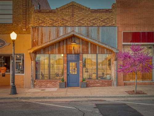 A rustic storefront with a blue door, large windows, and a flowering tree in front, set on a charming street.