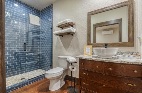 Modern bathroom featuring a glass shower, blue tiled wall, wooden vanity, and decorative shelves with towels.