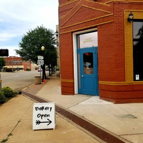 A bakery with a blue door and an open sign, located on a street with trees and buildings in the background.