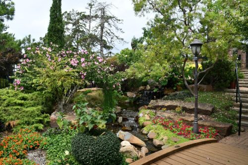 Lush garden with colorful flowers, a small stream, and a stone pathway, surrounded by trees and greenery.