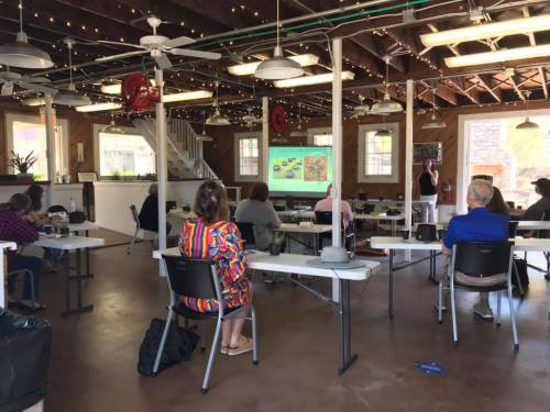 A classroom setting with attendees seated at tables, watching a presentation on a screen in a well-lit space.