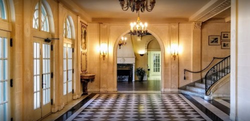 Elegant foyer with arched doorways, chandelier, and checkered floor leading to a staircase and fireplace.