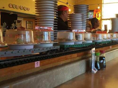 Conveyor belt sushi restaurant with stacked plates and diners in the background.