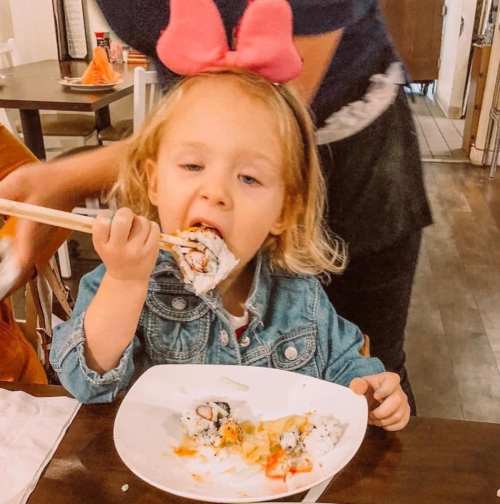 A young girl in a denim jacket and pink bow enjoys sushi with chopsticks at a dining table.