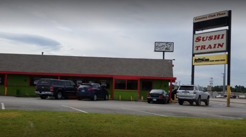 Exterior view of Sushi Train restaurant with a sign, parked cars, and a cloudy sky in the background.