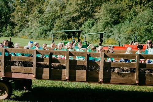 A group of children in green shirts sitting on a hay wagon, smiling and waving in a sunny outdoor setting.