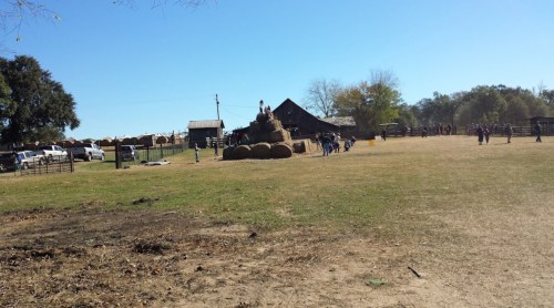 A sunny farm scene with people near hay bales and a barn, surrounded by open fields and trees.