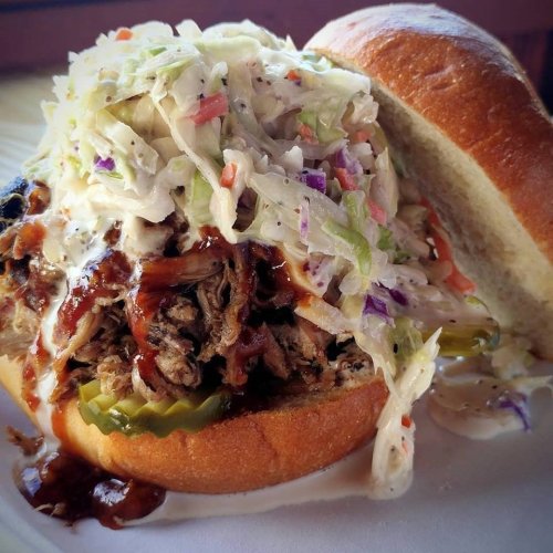 A pulled pork sandwich topped with coleslaw and pickles, served on a soft bun with barbecue sauce.