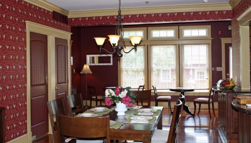 Cozy dining area with a wooden table, floral centerpiece, and warm red wallpaper, featuring large windows and a chandelier.