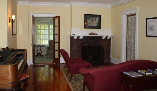 Cozy living room with a piano, two red chairs, a fireplace, and a view of a sunlit room through an open door.