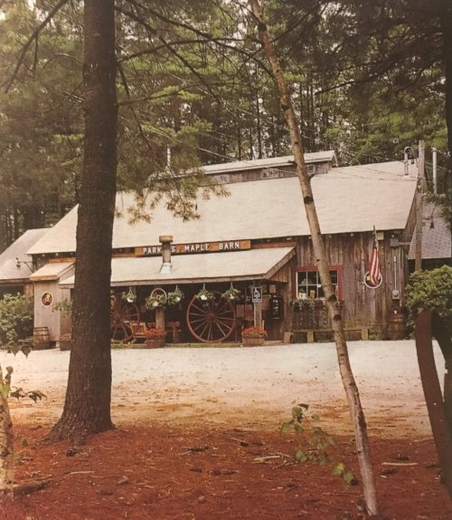 A rustic barn surrounded by trees, featuring a sign, flower pots, and a wagon wheel in front.