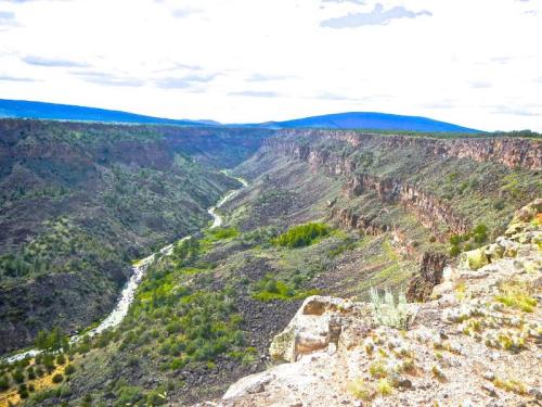 A scenic view of a deep canyon with a winding river, surrounded by rocky cliffs and green vegetation under a cloudy sky.