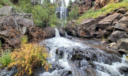 A serene waterfall cascades over rocks, surrounded by lush greenery and autumn foliage.