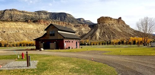 A rustic building near a grassy area, with mountains and a rocky cliff in the background under a clear sky.