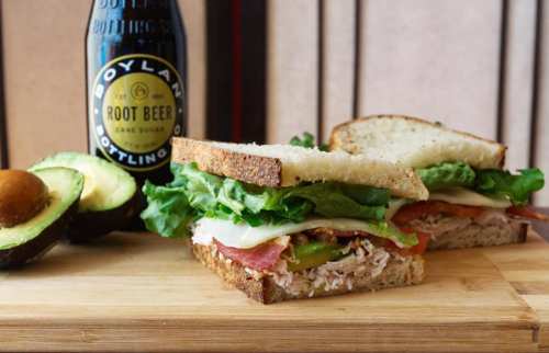 A close-up of a sandwich with lettuce, tomato, and turkey, alongside a bottle of root beer and an avocado on a wooden board.