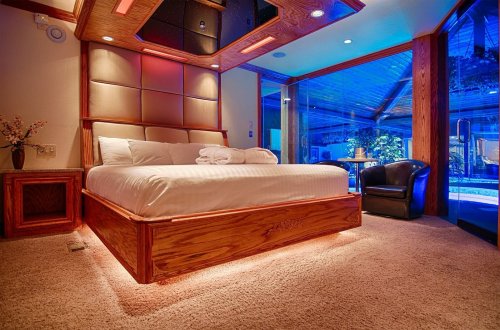 A modern bedroom with a large bed, warm lighting, and a view of a blue-lit outdoor area through glass doors.