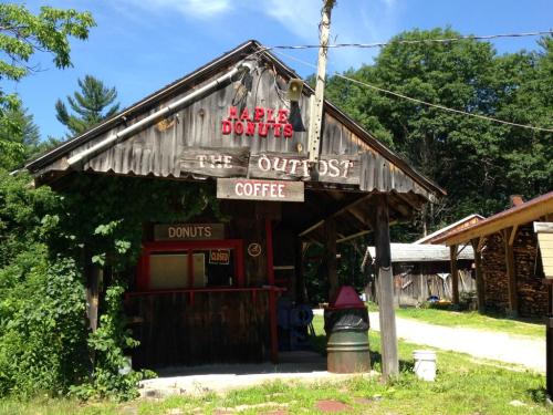 A rustic wooden building with a sign reading "Maple Donuts" and "The Outpost," surrounded by greenery and a clear blue sky.