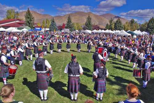 A large crowd watches a bagpipe band performing outdoors, surrounded by trees and tents under a blue sky.