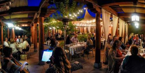 A lively outdoor restaurant scene at night, with diners enjoying meals under string lights and a musician playing nearby.