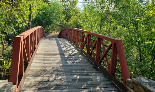 A wooden bridge with a red metal railing spans a path surrounded by lush green trees and foliage.