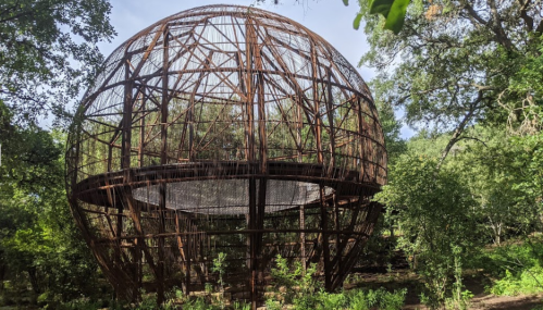 A large, rusted spherical structure surrounded by lush greenery and trees.