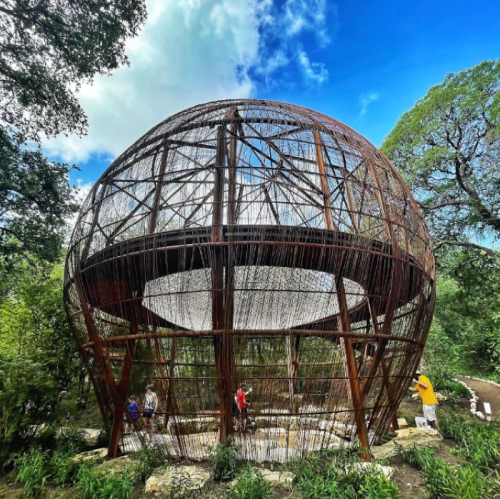 A large, spherical structure made of woven branches, surrounded by greenery and blue skies. People are walking nearby.
