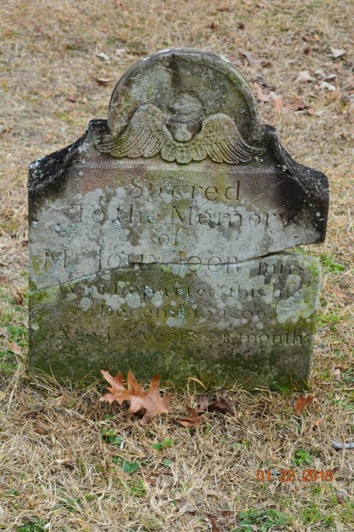 Weathered gravestone with an angel motif, inscribed in memory of a person, partially obscured by moss and dirt.