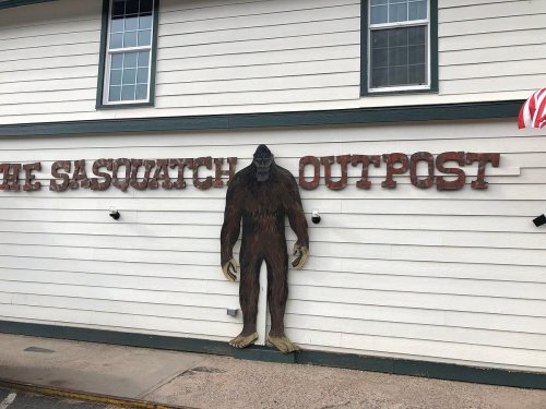 A large Sasquatch figure stands beside the "Sasquatch Outpost" sign on a white building.