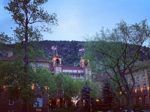 Historic hotel surrounded by trees, with flags flying and mountains in the background, illuminated at dusk.