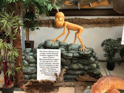 A sculpture of the Dover Demon, an orange creature, perched on rocks with a sign about its sighting in Dover, Massachusetts.